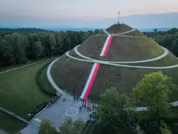 Pomnik w kształcie kopca z biało-czerwonymi wstęgami i flagami, otoczony zielenią.