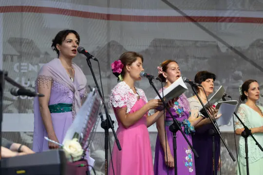 Grupa kobiet w kolorowych strojach śpiewa na scenie, trzymając nuty.