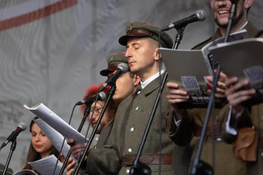 Mężczyzna w mundurze wojskowym recytuje tekst, wokół niego inni wykonawcy z mikrofonami.