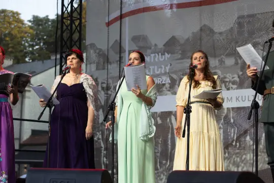 Cztery kobiety w strojach ludowych śpiewają na scenie, trzymając kartki z tekstem.