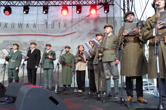 Grupa osób w mundurach śpiewa na scenie podczas wydarzenia upamiętniającego.