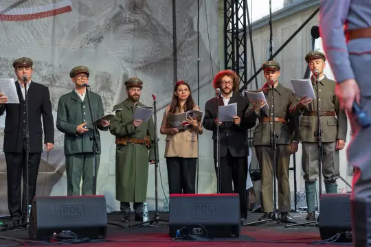 Grupa osób w strojach historycznych śpiewa na scenie, trzymając teksty utworów.