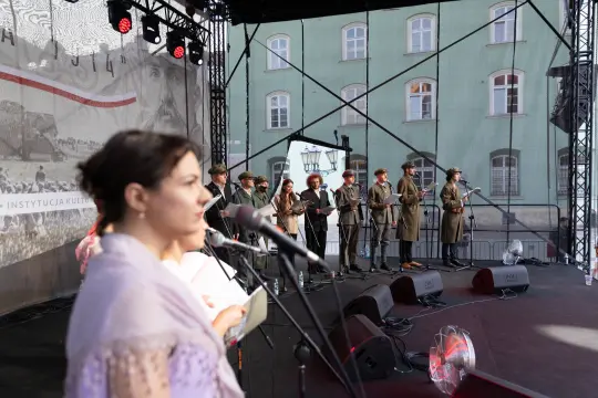 Występ artystyczny na scenie z uczestnikami w mundurach i mikrofonami.