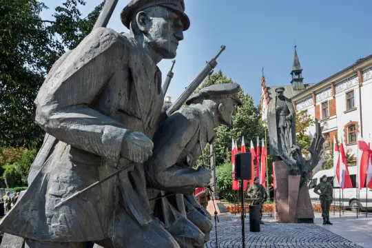 Pomnik żołnierzy z bronią, flagi narodowe w tle, symbolika patriotyczna.