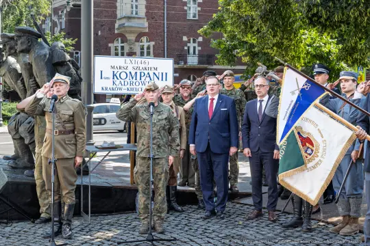 Uroczystość z okazji marszu, uczestnicy w mundurach, flagi i pomnik w tle.