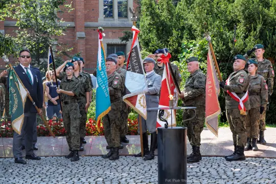 Uroczystość z udziałem żołnierzy i sztandarów, w tle kwiaty i budynki.