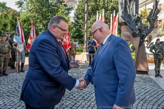 Dwaj mężczyźni w garniturach podają sobie ręce w otoczeniu flag i żołnierzy.