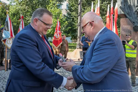 Dwaj mężczyźni wymieniają uściski dłoni, w tle flagi i żołnierze.