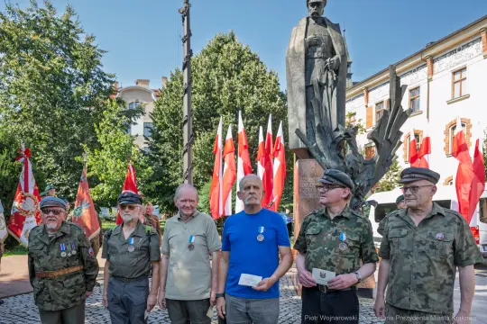 Grupa mężczyzn w mundurach wojskowych stoi przed pomnikiem, w tle flagi Polski.