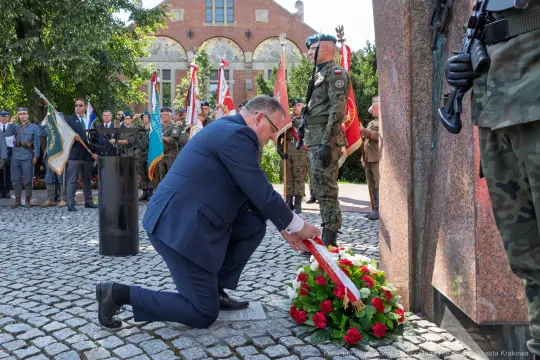 Mężczyzna składa wieniec przy pomniku, w tle żołnierze i flagi.