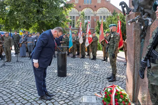 Uroczystość złożenia hołdu przy pomniku, żołnierze i delegacje z flagami.