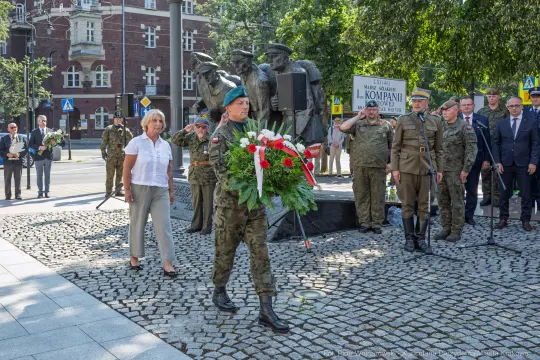 Żołnierz składa wieniec w hołdzie, obok grupy osób i pomnika.