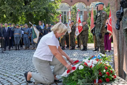 Kobieta składa kwiaty przy pomniku, w tle żołnierze i uczestnicy uroczystości.