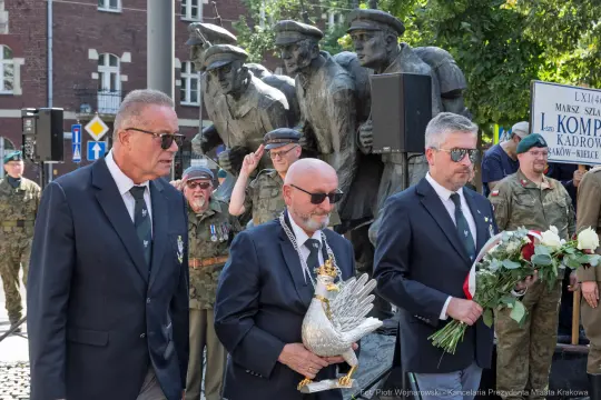 Mężczyźni w garniturach niosący symboliczne odznaczenie podczas uroczystości upamiętniającej.
