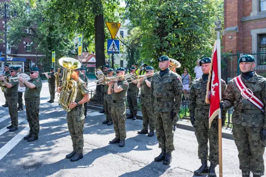Orkiestra wojskowa w mundurach, grająca na ulicy podczas uroczystości.