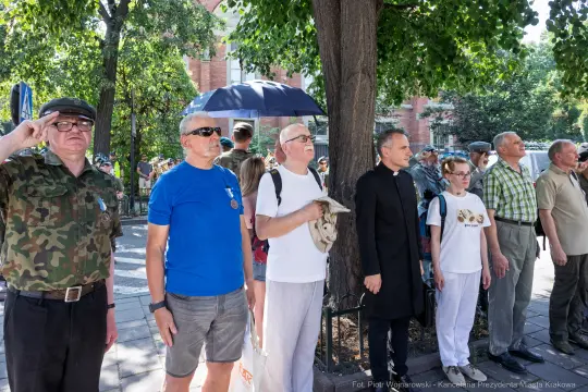 Grupa ludzi w różnych strojach, niektórzy w mundurach, stojących w kolejce pod drzewem.