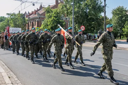 Żołnierze w mundurach wojskowych maszerują w defiladzie, trzymając flagi.