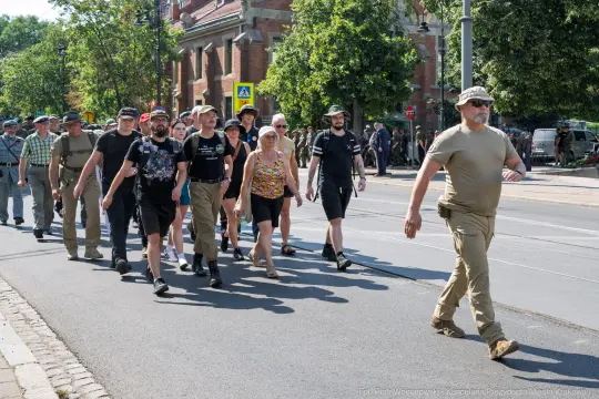Grupa ludzi idąca ulicą, część z nich w mundurach, w tle budynki i drzewa.