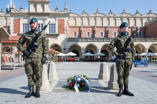 Dwóch żołnierzy w mundurach wojskowych stoi na placu, obok leży wieniec kwiatów.
