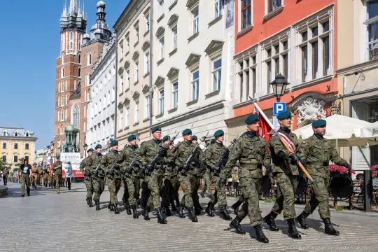 Żołnierze w mundurach maszerują ulicą, trzymając flagi i sztandary.