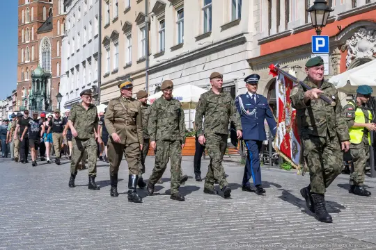 Grupa żołnierzy i oficerów w mundurach idąca ulicą, obok tłumu ludzi i kamienic.