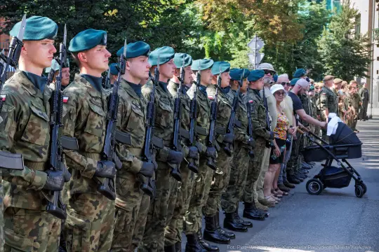 Żołnierze w mundurach stoją w szeregu, obok nich cywile z dzieckiem w wózku.