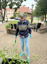 Osoba pielęgnująca rośliny w parku, w rękach trzyma łopatkę, ubrana w niebieskie rękawice.