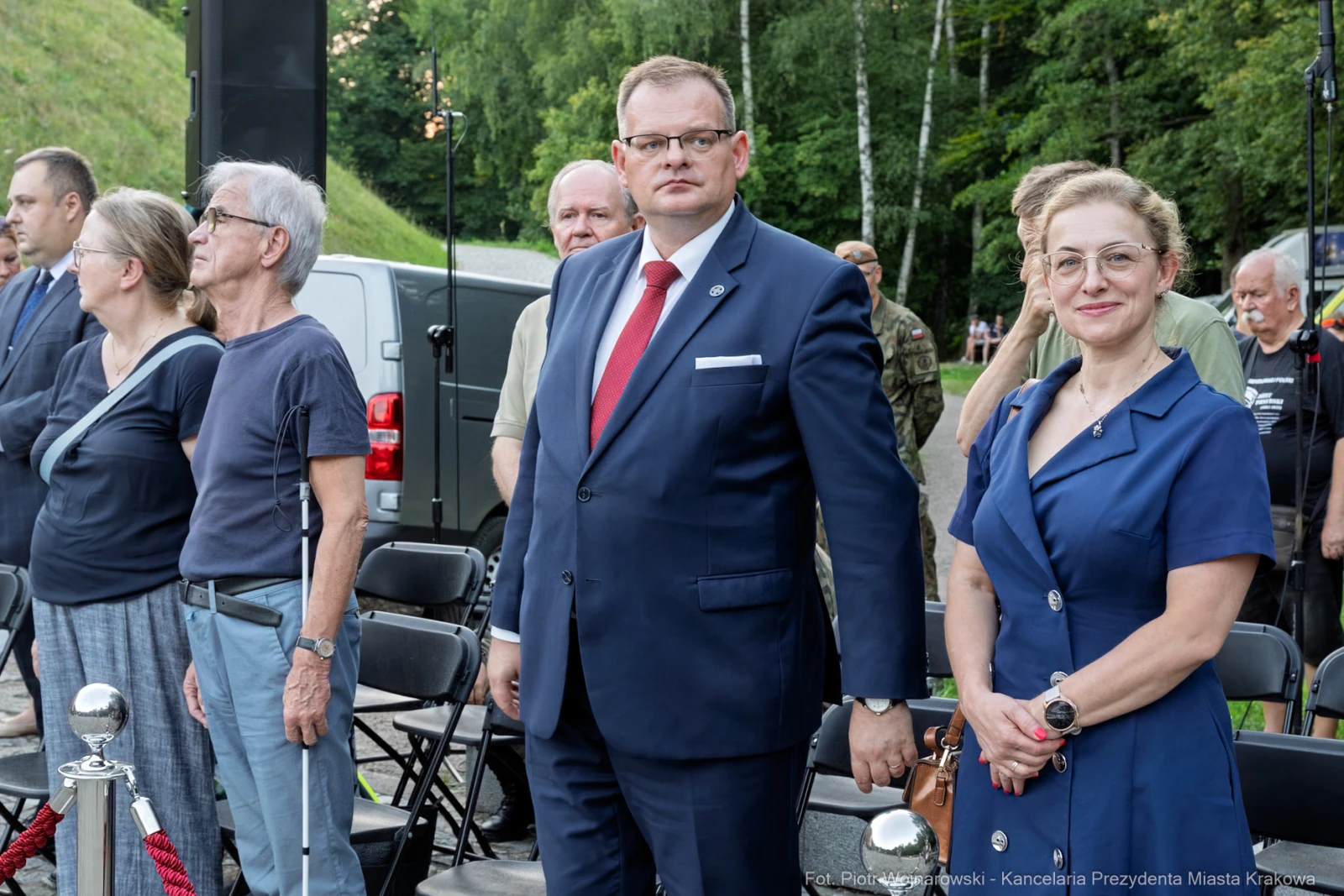 Grupa ludzi stojąca w kolejce, mężczyzna w garniturze i kobieta w niebieskiej sukience.  Autor: Piotr Wojnarowski