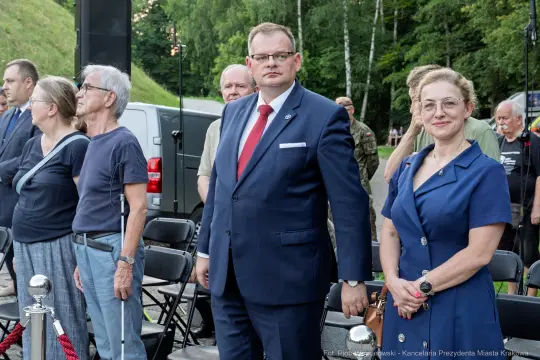 Grupa ludzi stojąca w kolejce, mężczyzna w garniturze i kobieta w niebieskiej sukience.