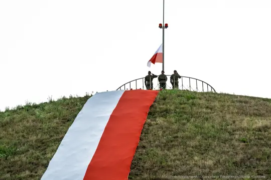 Żołnierze przy polskiej fladze na wzgórzu, obok rozwinięta flaga narodowa.