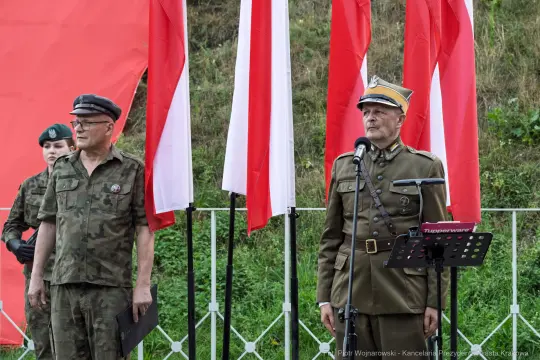 Dwaj mężczyźni w mundurach wojskowych stoją przed flagami Polski, jeden z nich przemawia.