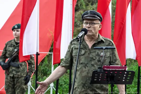 Mężczyzna w mundurze wojskowym przemawia do mikrofonu, obok stoi żołnierz. W tle flagi.