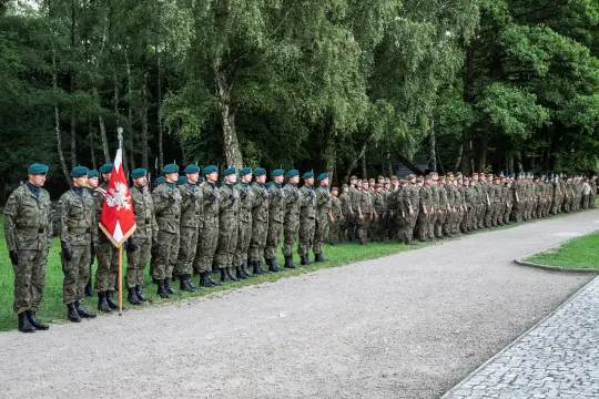 Żołnierze w mundurach stoją w szeregu, trzymając flagę. Drzewa w tle.