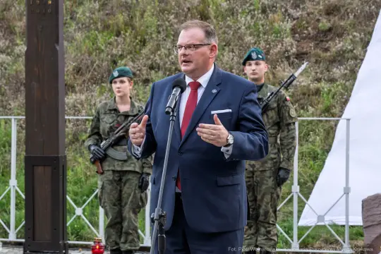 Mężczyzna w garniturze przemawia do mikrofonu, w tle żołnierze w mundurach.