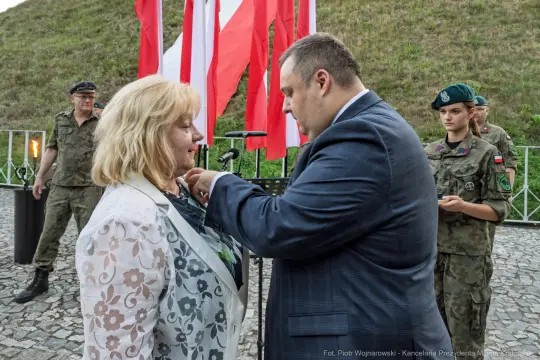 Ceremonia wręczenia odznaczenia, kobieta odbiera medal od mężczyzny w garniturze, w tle flagi i żołnierze.