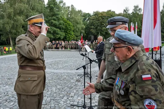 Ceremonia wojskowa z żołnierzami i weteranami, flagi Polski w tle.