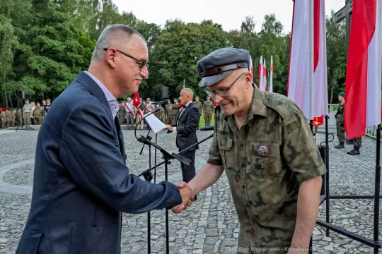 Uścisk dłoni dwóch mężczyzn w mundurach wojskowych, w tle flagi i przemawiający mężczyzna.