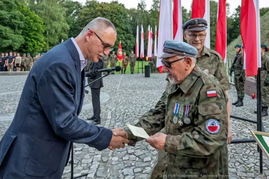 Wręczenie odznaczeń wojskowych podczas ceremonii z flagami Polski w tle.