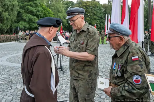 Wręczenie odznaczenia weteranowi przez oficera w mundurze, w tle flagi i zgromadzeni ludzie.