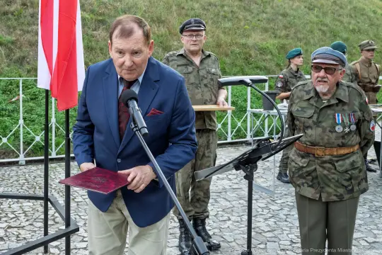 Mężczyzna w garniturze przemawia przy flagach, w tle żołnierze w mundurach.