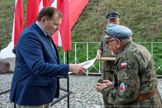 Wręczenie odznaczenia przez mężczyznę w garniturze weteranowi w mundurze. W tle flagi.