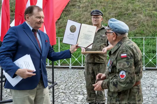 Wręczenie odznaczenia z okazji 35-lecia, mężczyźni w mundurach i garniturze, flagi w tle.