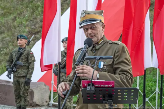 Mężczyzna w mundurze wojskowym przemawia do mikrofonu, w tle flagi Polski i żołnierze.