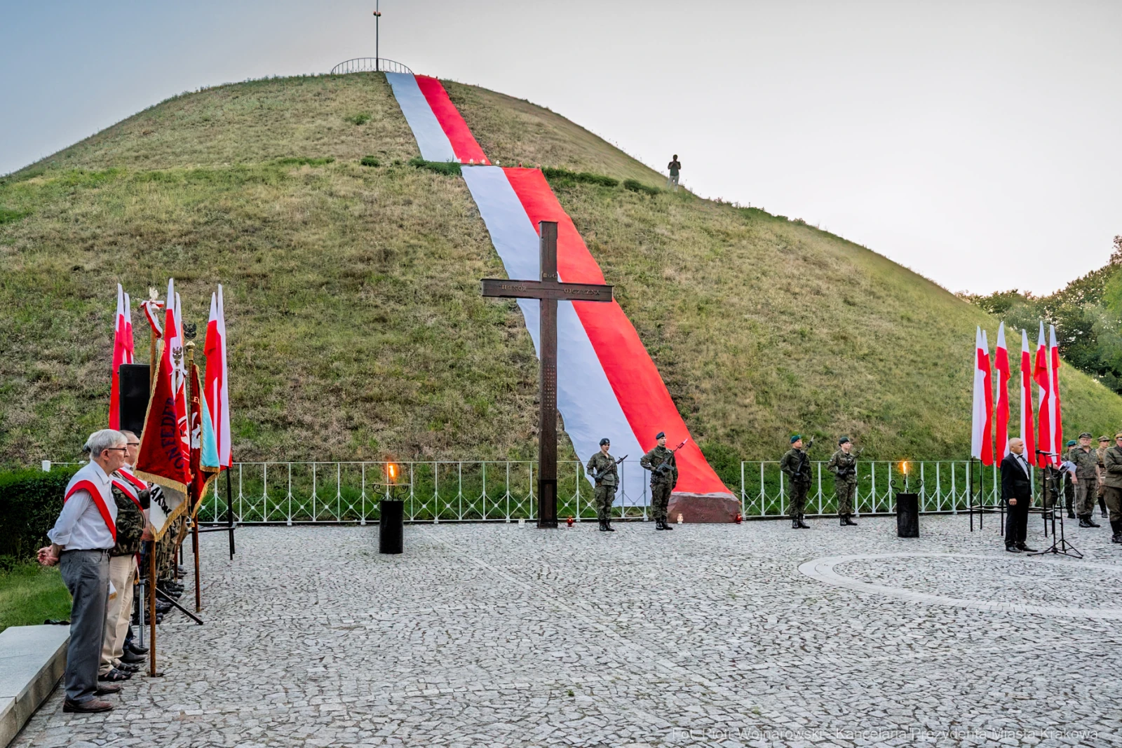 Uroczystość z flagami i krzyżem przed wzgórzem z biało-czerwoną dekoracją.  Autor: Piotr Wojnarowski