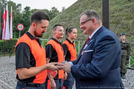 Uroczystość wręczenia odznaczeń przez przedstawiciela władz. Czerwone kamizelki, flagi Polski w tle.