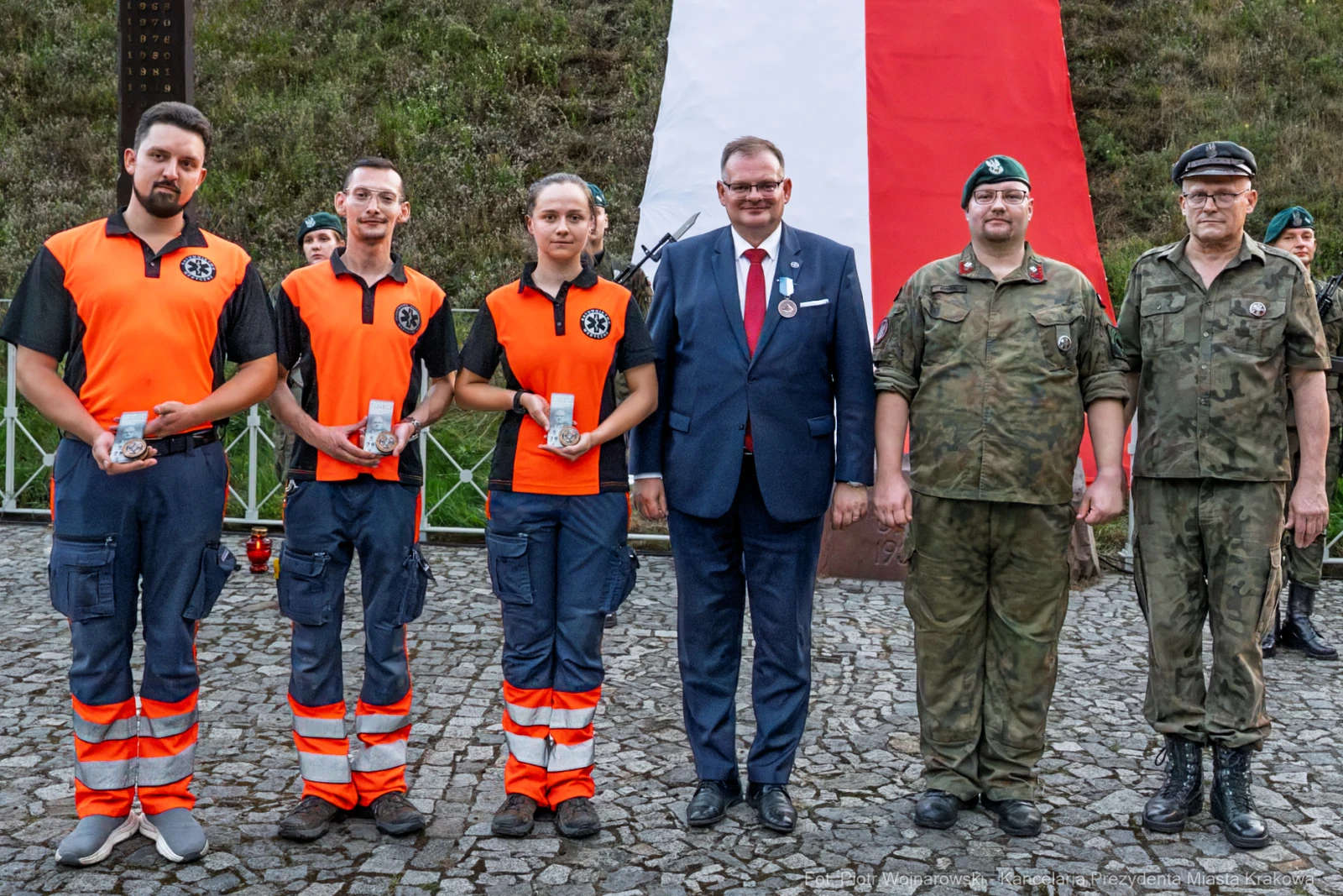 Cztery osoby w pomarańczowych i granatowych strojach oraz trzech mężczyzn w mundurach, stojących przed flagą.  Autor: Piotr Wojnarowski