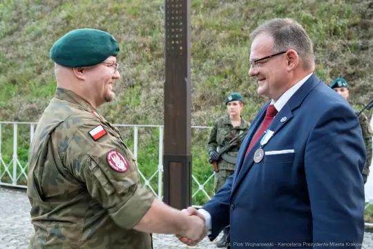 Dwaj mężczyźni w mundurze wojskowym i garniturze uścisną ręce podczas ceremonii.