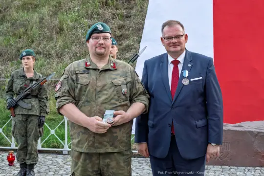 Mężczyzna w mundurze wojskowym trzyma medal, obok stoi mężczyzna w garniturze. W tle flaga.