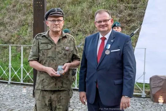 Dwaj mężczyźni w mundurze wojskowym i garniturze, jeden z medalem, podczas ceremonii.