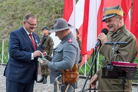 Uroczystość z udziałem mężczyzn w mundurach, flagi Polski w tle.